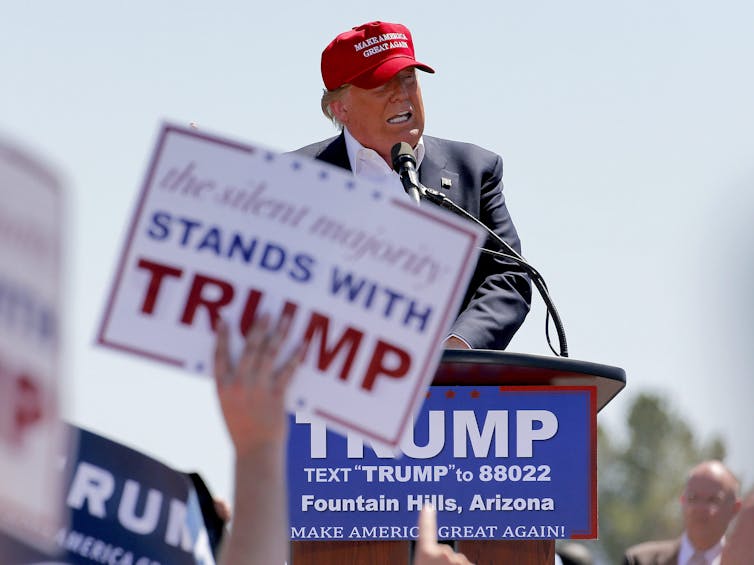 A man in a red ball cap speaks into a microphone. In front of him a woman holds up a sign that reads The Silent Majority Stands With Trump.