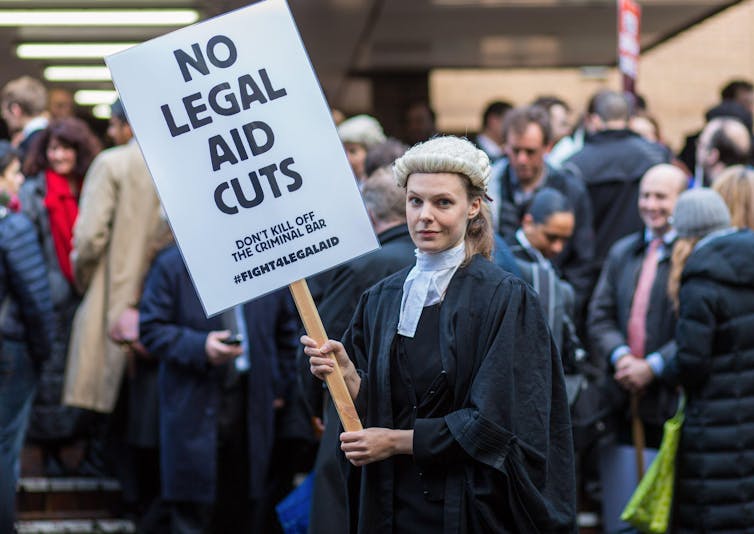 A female barrister in a white wig holds a sign reading 'no legal aid cuts'.
