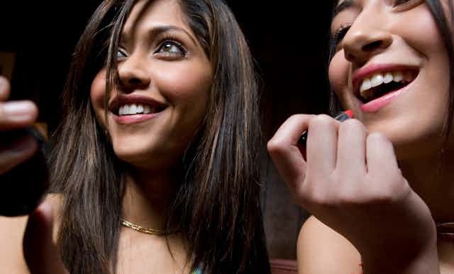 Two young women put on makeup while holding a mirror. They're smiling and look like they're ready to go to a party.