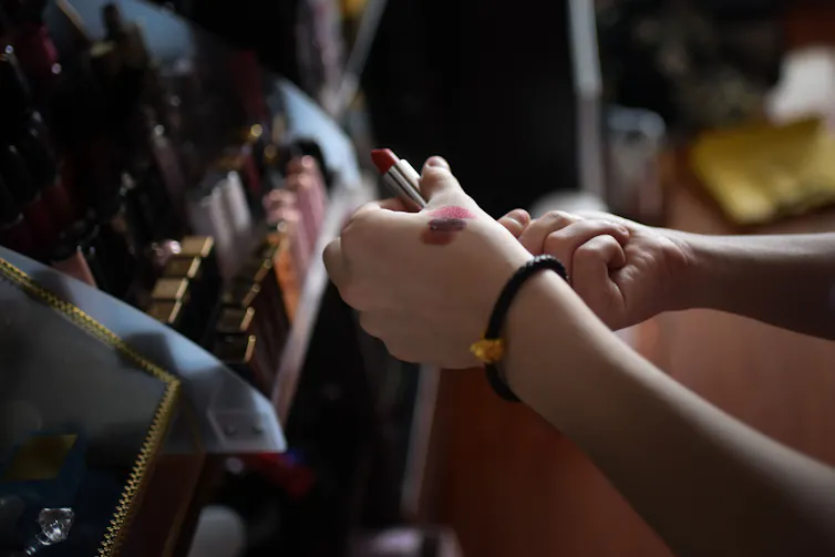 Les mains d'une personne testent des couleurs de rouge à lèvres dans un magasin, devant un comptoir rempli de cosmétiques.