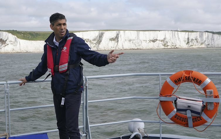 A man in a lifejacket stands on a boat in front of white cliffs