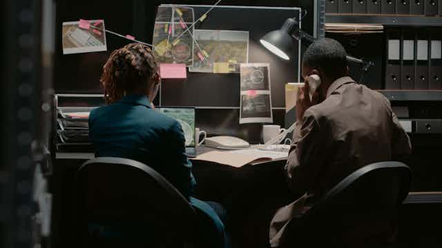 A woman staring at a laptop screen and a man holding a corded phone seated in front of a black noticeboard with pinned images. A black lamp illuminates their desk.