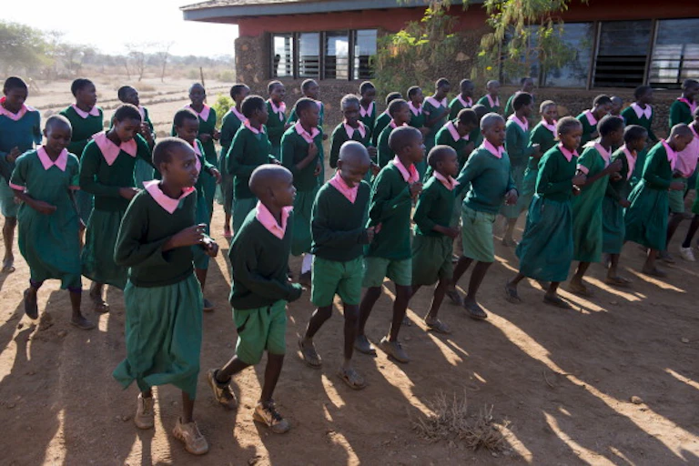 Boys and girls dressed in a school uniform of green dresses with pink collars, and green sweaters and green shorts with pink shirts walking in three rows in front of a building