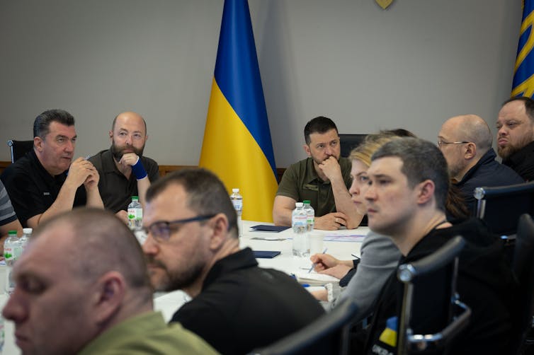 Volodymyr Zelensky sits at the head of a table of people in dicussion.