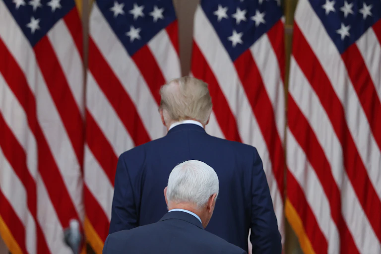 The backs of two men wearing blue suits are seen in front of a row of American flags.
