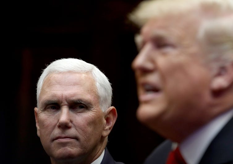 A man with white hair looks to his side at a man with an open mouth and light white hair who is speaking.