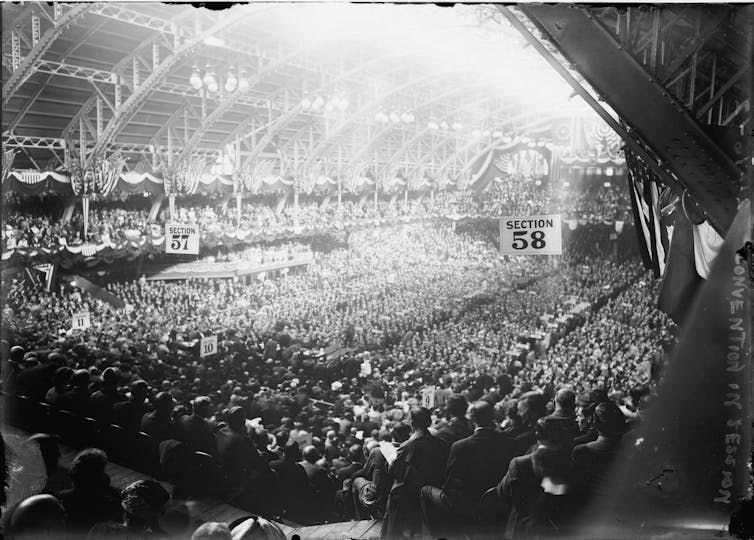 A black and white photo shows a large room filled with people, in a stadium like setting.