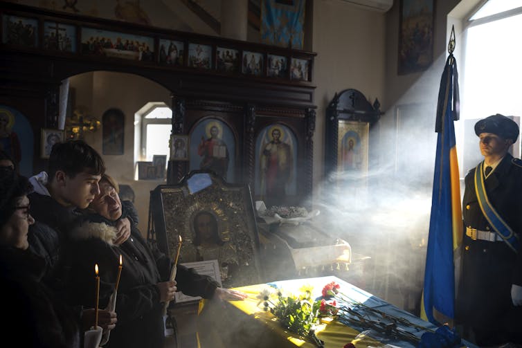 A sunbeam illuminates a coffin covered in flowers as a mother and young man stand in the shadow. The woman is crying.