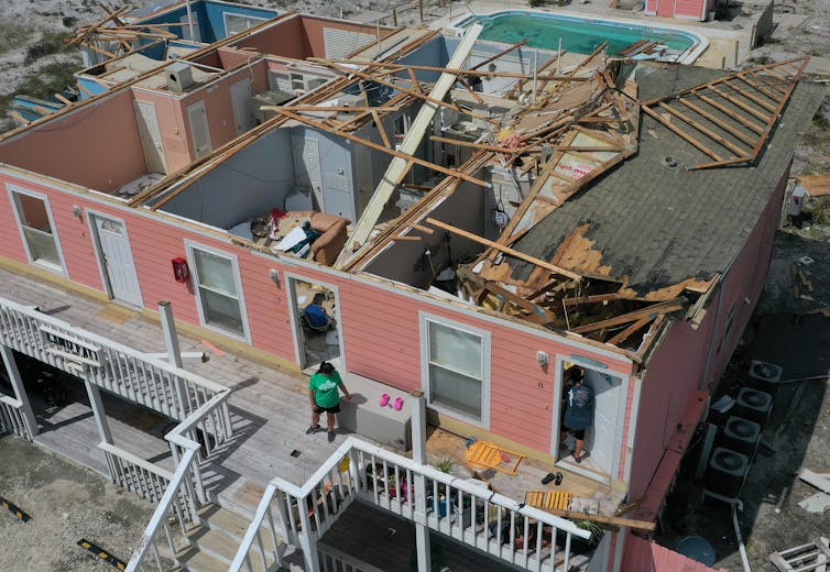 A one-story apartment building on stilts with the roof torn off after Hurricane Sally. Pink beach shoes and folded beach chairs sit on a porch.