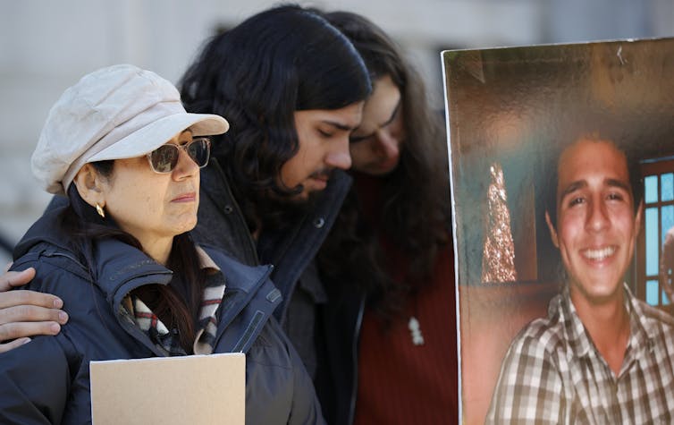 Three people looking sad hold up a photo of a smiling man.