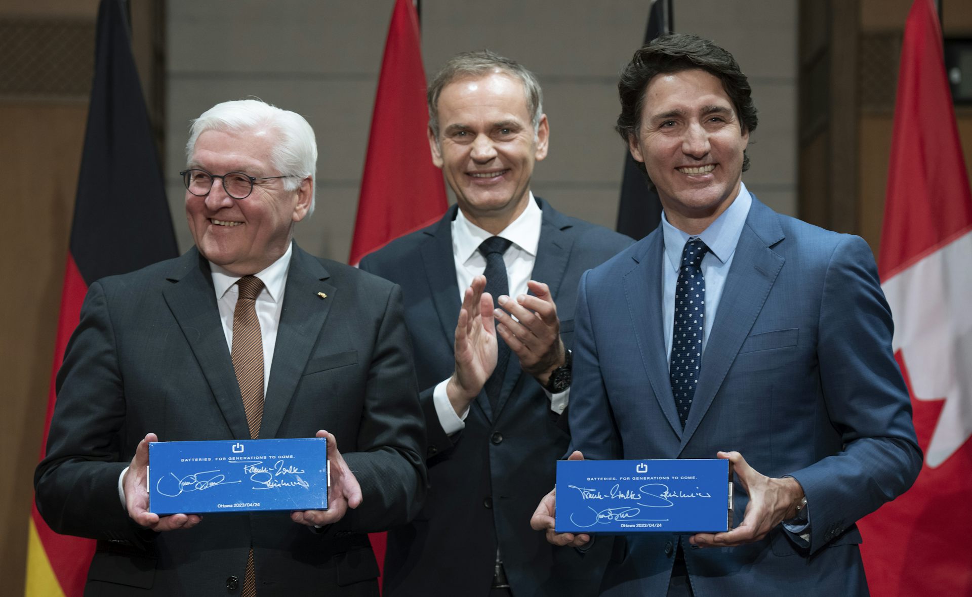 Three men, two holding batteries, smile for the cameras. A row of Canadian flags is behind them.