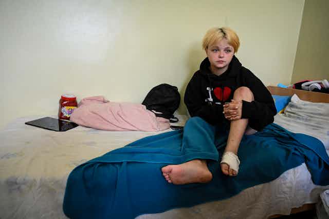 A young blond girl with a bandage wrapped around her left foot sits on her bed.