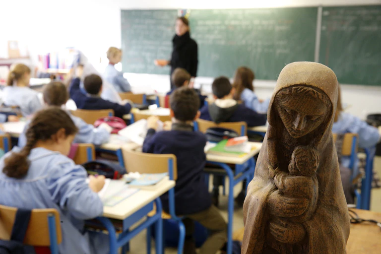 A blurry photo of young children sitting at desks in a classroom, with a wooden statue of a woman holding a baby in the foreground.