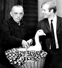 A serious looking older man is handing a goose in a basket to a cheerful younger man.