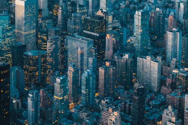 A aerial view of New York City at dusk as the lights are coming on in the buildings and the sky is reflected in the towers of glass.