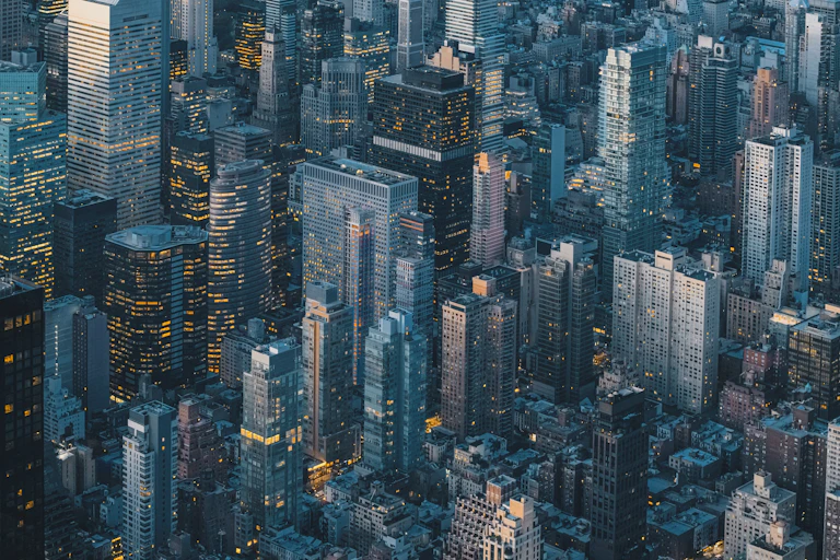 A aerial view of New York City at dusk as the lights are coming on in the buildings and the sky is reflected in the towers of glass.