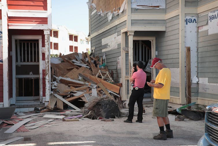 A man with a tape measure on his belt and camera looks at debris piles left between buildings after Hurricane Michael hit Florida. The siding of the building is also ripped off.