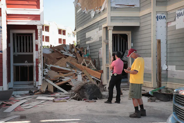 A man with a tape measure on his belt and camera looks at debris piles left between buildings after Hurricane Michael hit Florida. The siding of the building is also ripped off.