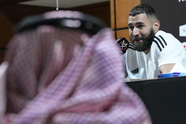 A man in a white soccer jersey speaks into a microphone in front of a man in traditional Arab headscarf.