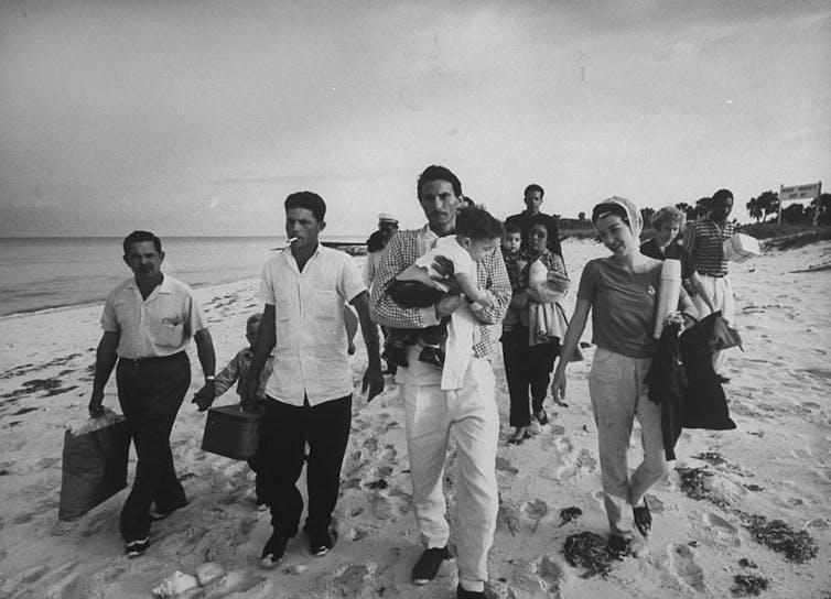 Fotografía en blanco y negro de cubanos paseando por la playa con equipaje y niños.