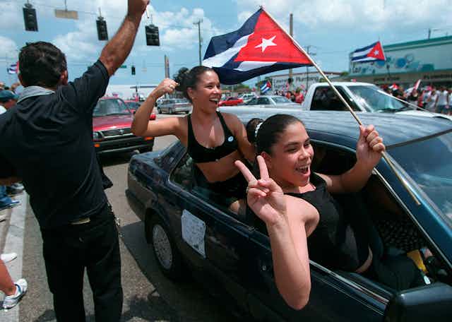 Two smiling young women hang out car windows while one waves a Cuban flag.