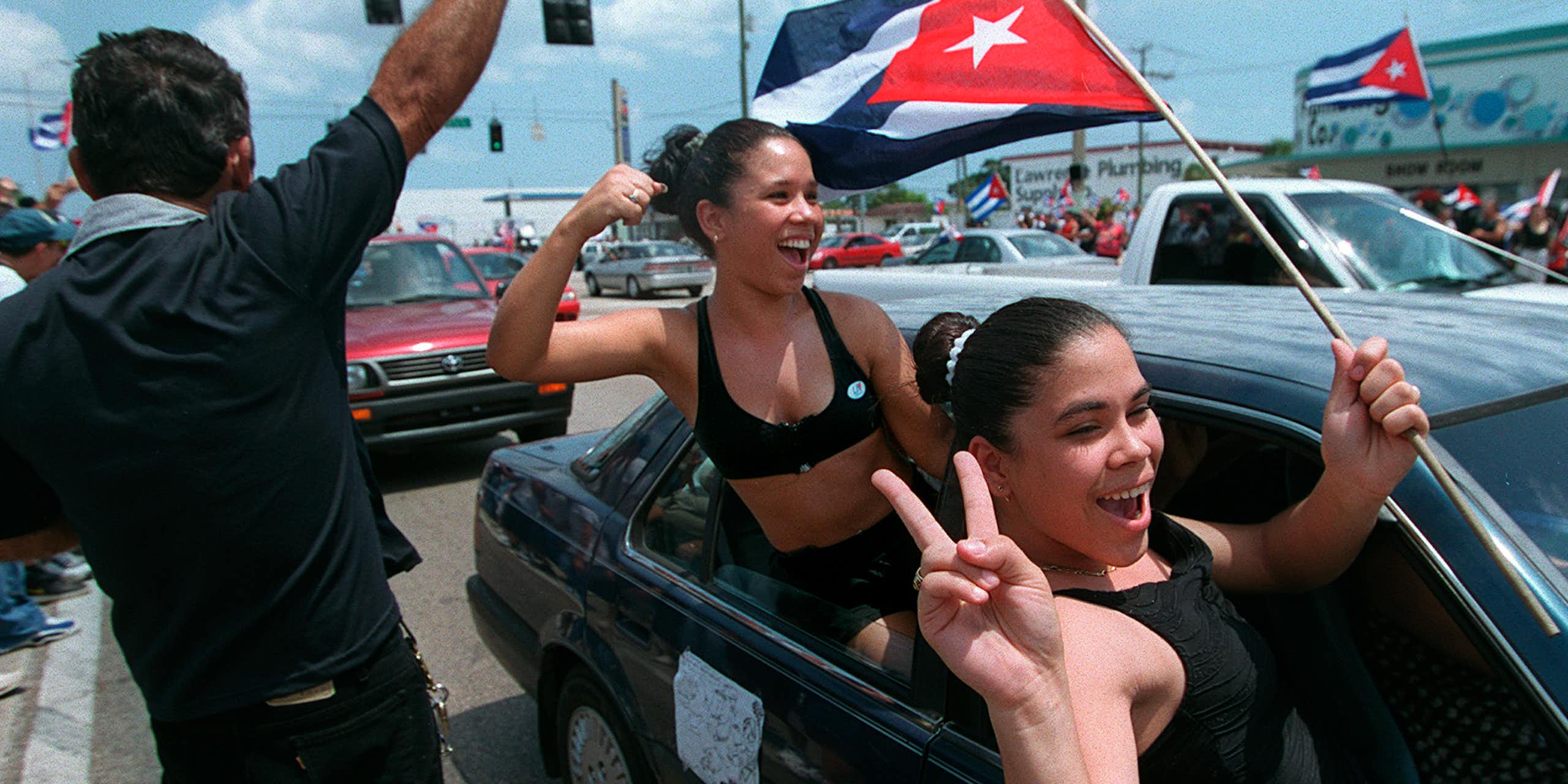 Two smiling young women hang out car windows while one waves a Cuban flag.