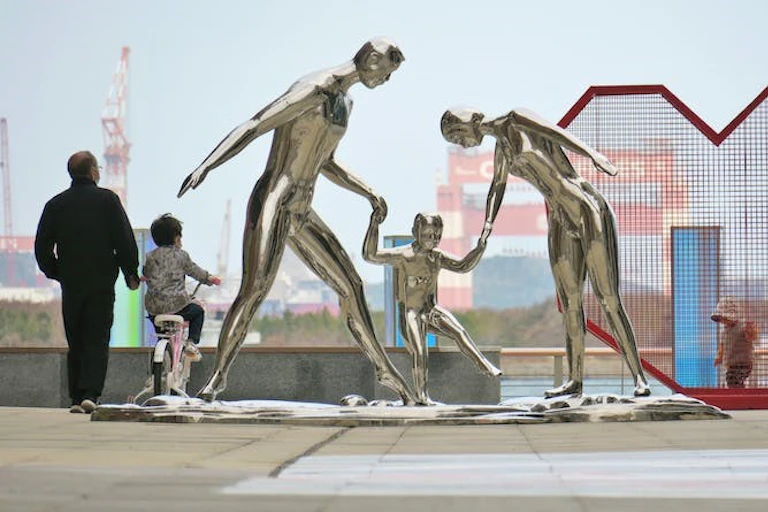 Un vieil homme guide un enfant à vélo près de statues représentant une famille.