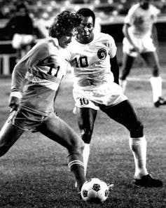 A man with long hair dribbles a ball past a player in a white shirt.