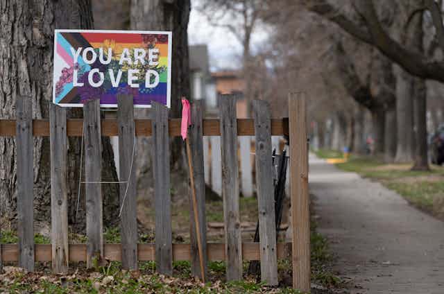 Poster of pride flag reading "You are loved" on top of a wooden fence beside a walkway