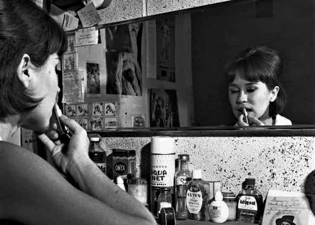Black and white photo of woman putting on lipstick in front of a mirror.