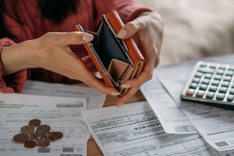 Woman holding empty purse open, surrounded by paperwork, a pile of coins, calculator.