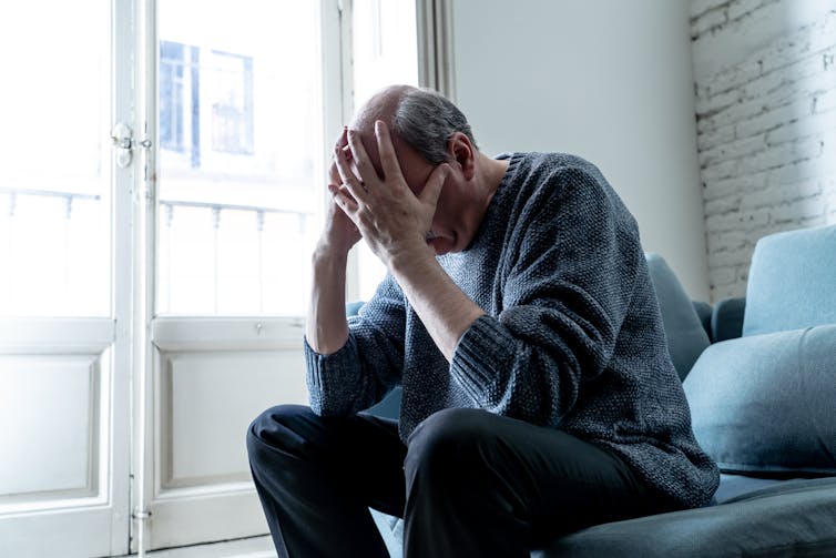 Older man sitting on sofa, head in hands, window, white walls.