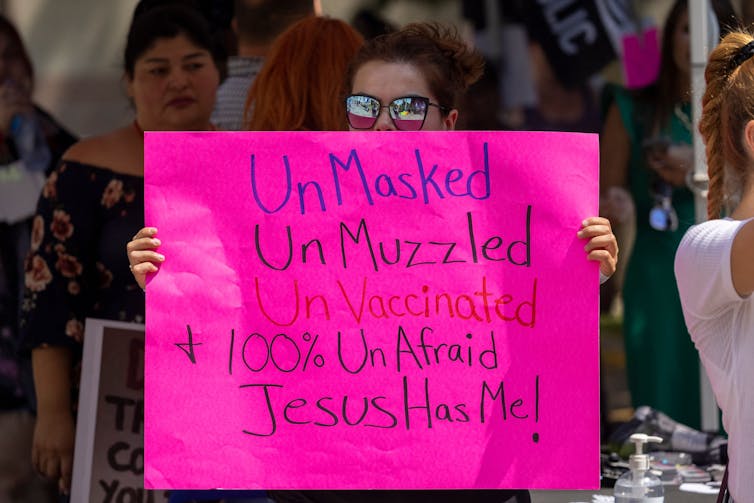 A woman wearing sunglasses holds a bright pink sign proclaiming her belief that she doesn't need to be vaccinated or wear a mask because Jesus will protect her from COVID-19, as anti-vaccination protesters pray nearby