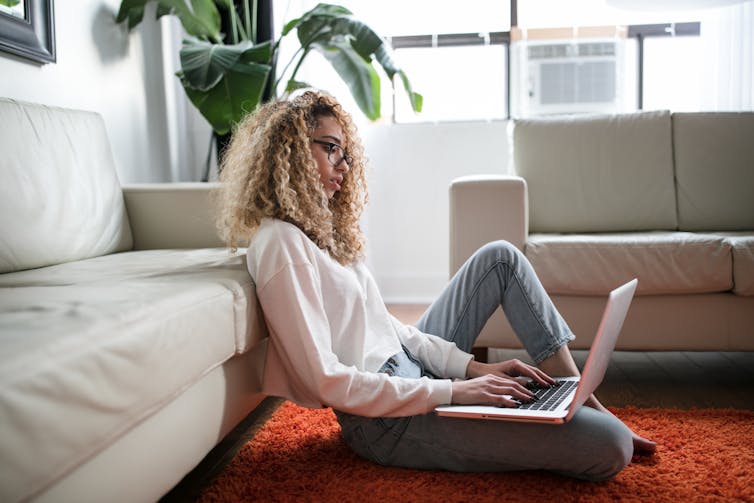 Woman sits on her longeroom floor, looking at her laptop