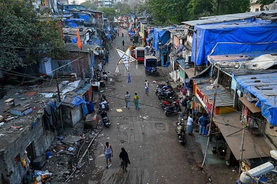 An aerial shot of men and women walking along a road littered with plastic, with makeshift houses made of plastic and iron sheets on either side of the road