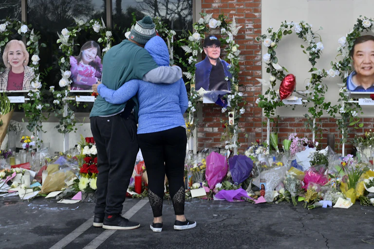 Two people stand hugging in front of a memorial wall covered with photographs and flowers.