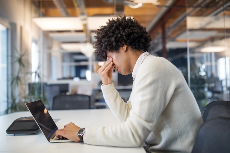 A man in an office looks down, away from his laptop, with a depressed demeanor.