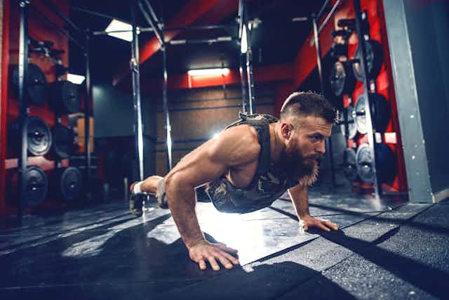 A man wearing a weighted vest does a push-up in a gym.