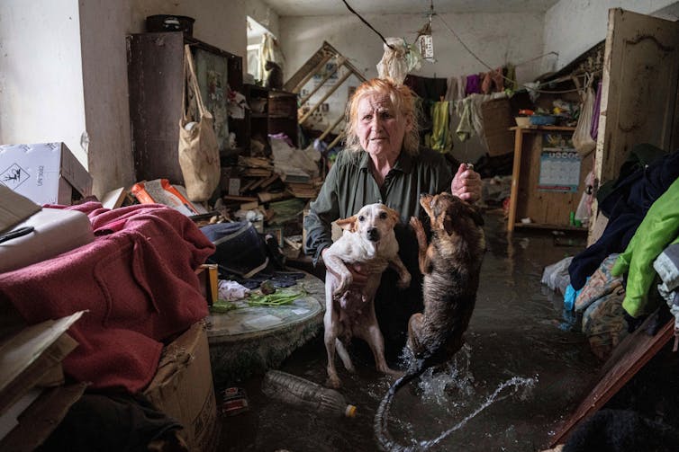A woman stands holding a dog and a cat in in a flooded room.