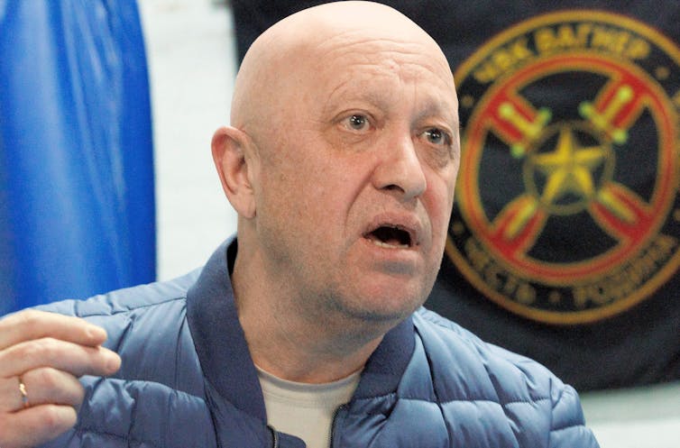 A bald man in a blue puffer jacket in front of a paramilitary insignia.