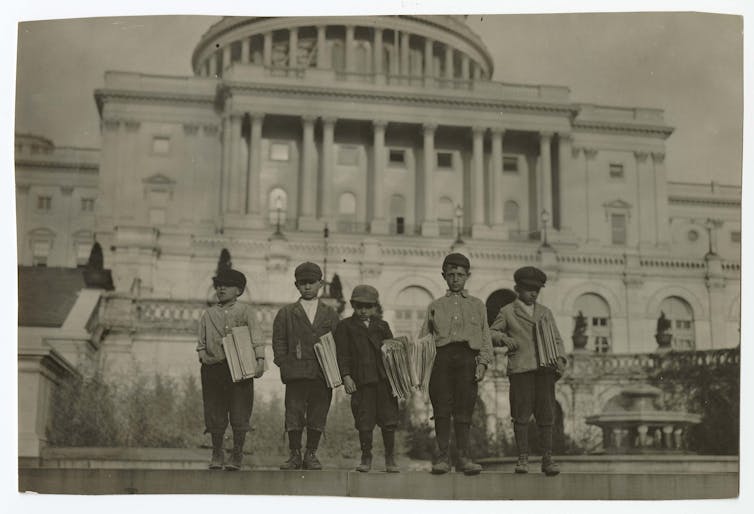 Five young boys wearing caps and holding newspapers in front of an imposing white building.