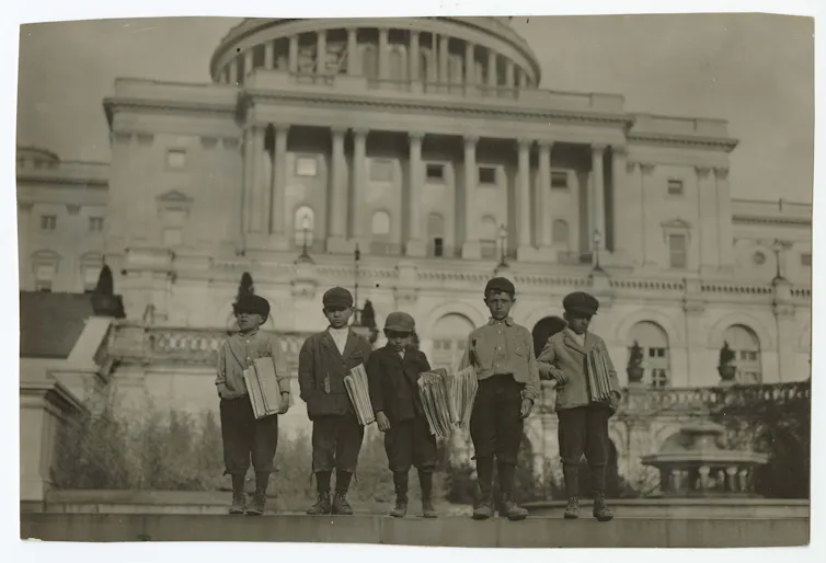 Five young boys wearing caps and holding newspapers in front of an imposing white building.