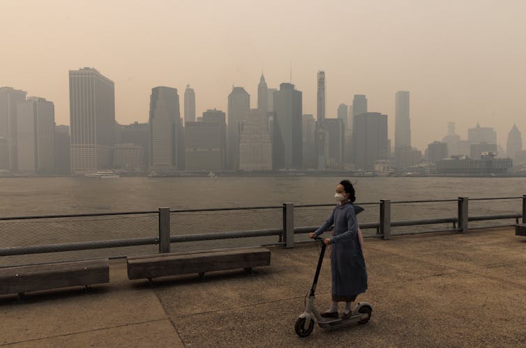 Uma mulher em uma scooter elétrica passa pelo horizonte de Nova York que é engolfado pela fumaça.