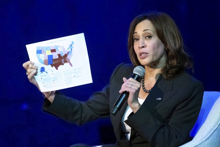 A woman in a suit speaks into a microphone while holding up a map of the United States