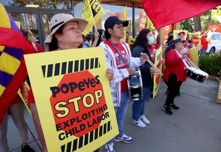 A group of women hold drums and signs reading 'Popeyes Stop Exploiting Child Labor.'