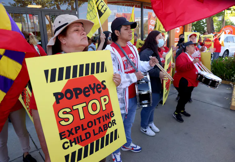 A group of women hold drums and signs reading 'Popeyes Stop Exploiting Child Labor.'