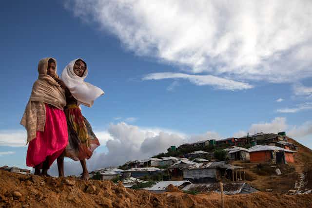 Two young girls with brown skin and headscarves embrace, while looking over a series of shacks on a hill. 