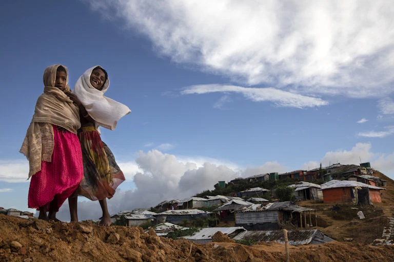 Two young girls with brown skin and headscarves embrace, while looking over a series of shacks on a hill.