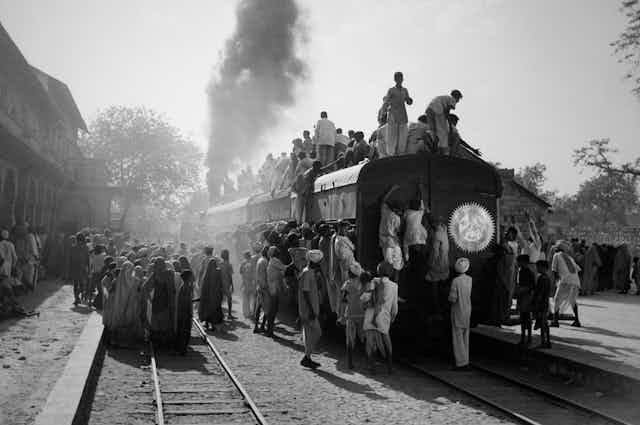 A plume of smoke emerges from the back of a train with people clambering all over the train.