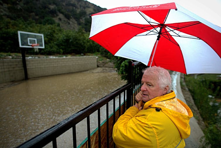 A man in a raincoat stands under a big umbrella watching his backyard fill with rainwater in California in 2023. California saw record rain from atmospheric rivers in early 2023.